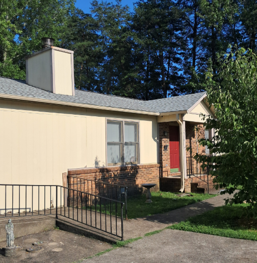 Single-story beige house with red front door, brick porch, and metal ramp in a sunny yard