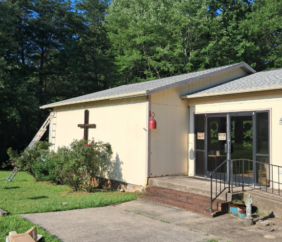 Small beige building with a cross on the wall, front steps, and a glass entrance surrounded by trees.