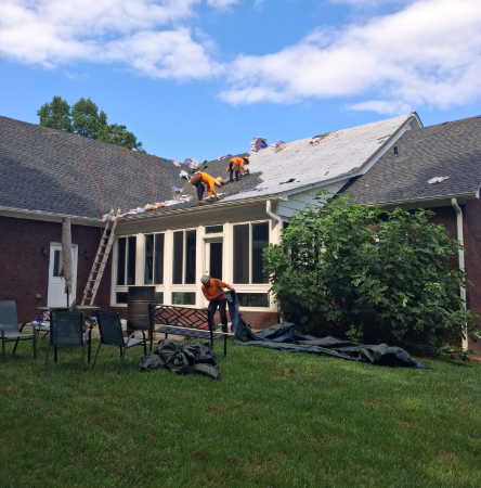 Workers replacing a house roof, with shingles removed and tarps on the lawn
