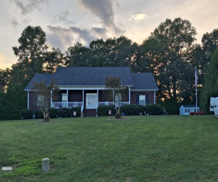 Single-story house with a dark roof and porch, set behind a wide grassy lawn at dusk.