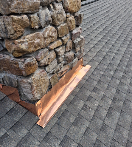 Stone chimney on a gray shingled roof with copper flashing at the base.