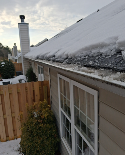 Snow-covered roof above a window beside a wooden fence and chimney in a residential yard.
