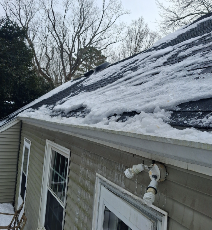 Snow-covered shingled roof beside beige siding and windows on a house