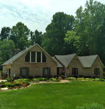 Brick house with a manicured green lawn, flower beds, and tall trees under a partly cloudy sky