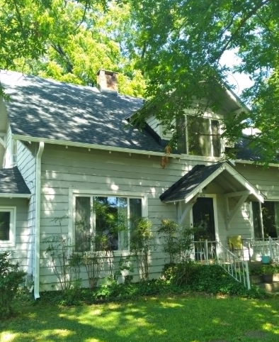 Light green house with dark roof and white trim, partly shaded by leafy trees and sunlight.