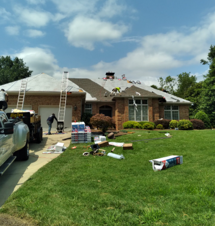 Workers on a brick house roof with ladders, tools, and roofing materials on the lawn under a blue sky