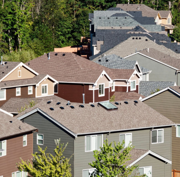 Aerial view of suburban houses with gray and brown roofs beside a wooded area.
