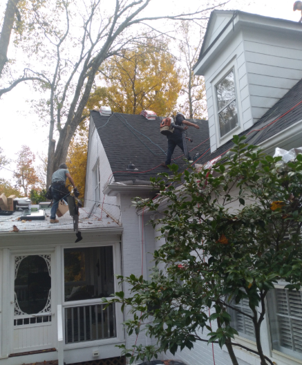 Two workers on a house roof beside a white siding home and tree branches