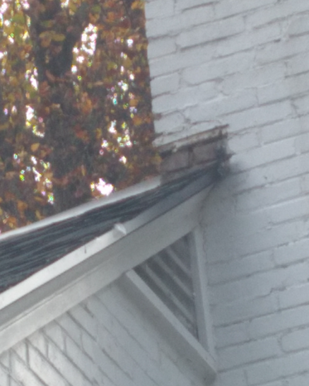 White house roof corner with damaged shingles near a chimney, framed by autumn trees.