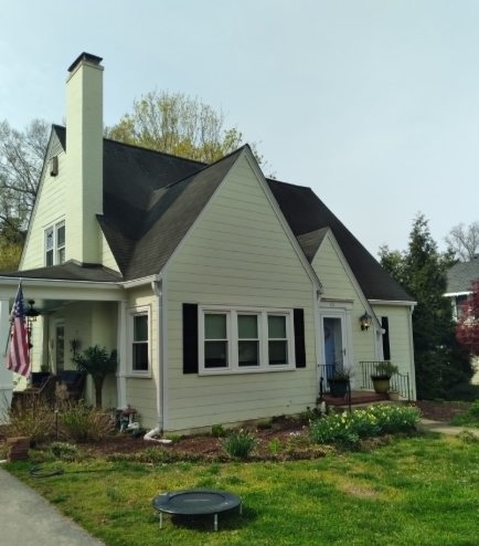 Cream-colored house with dark roof, chimney, and front garden on a tree-lined street