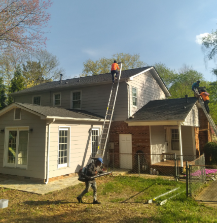 Workers on ladders installing a roof on a house with a brick chimney and white siding.