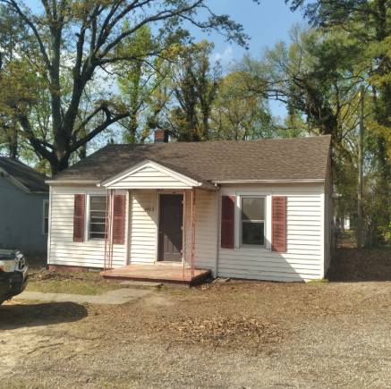Small white house with a front porch, brown roof, and trees in the yard under a clear sky