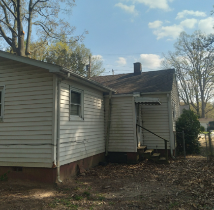Side view of a white house with a small porch, red foundation, and trees in a yard