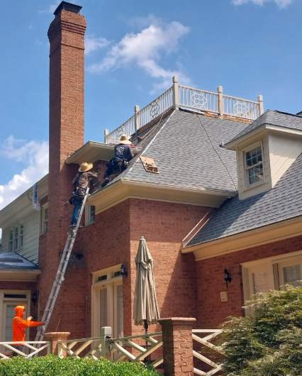 Workers on a ladder repairing a steep gray roof beside a red brick chimney and house.