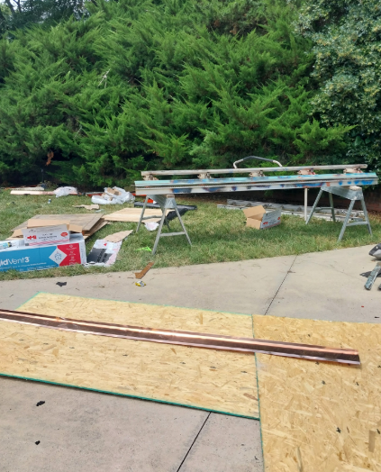 Outdoor work area with plywood sheets, sawhorses, and tools beside dense green bushes.