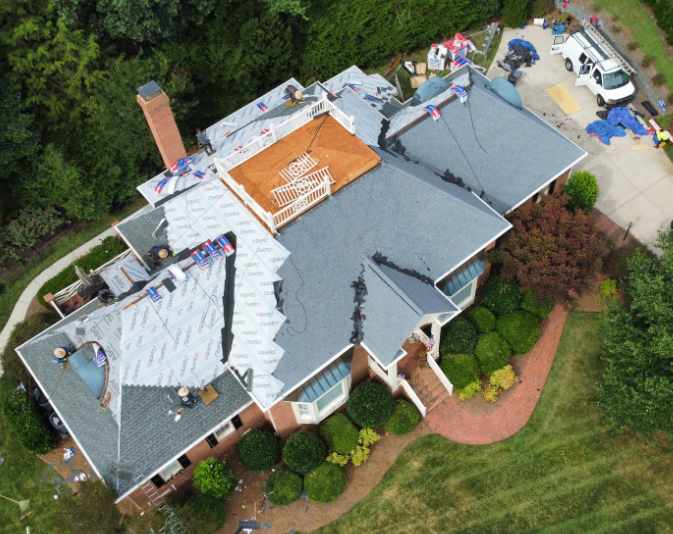 Aerial view of a house with roof work in progress, showing white underlayment, workers, and roofing materials.