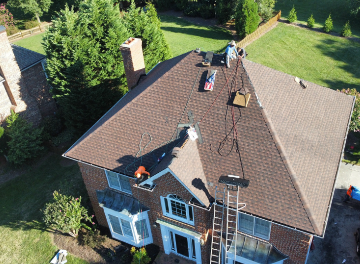 Aerial view of workers repairing a brown shingle roof on a brick house with a ladder and tools.