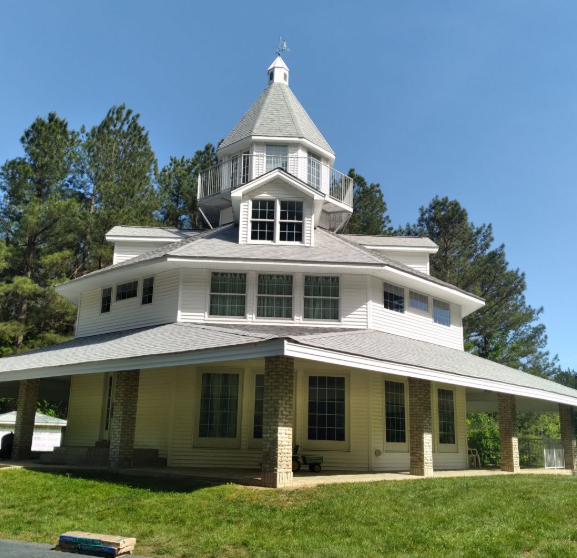 White Victorian-style house with wraparound porch and tower, surrounded by trees and grass.