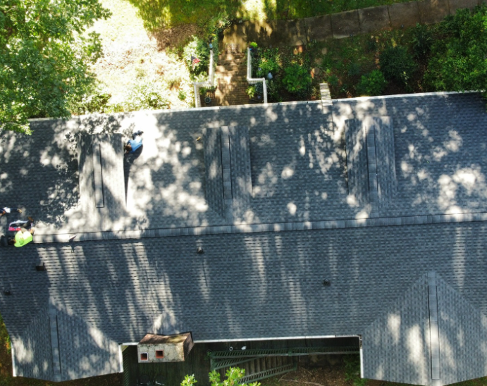 Two workers on a gray shingled roof beside a wall and trees, seen from above.