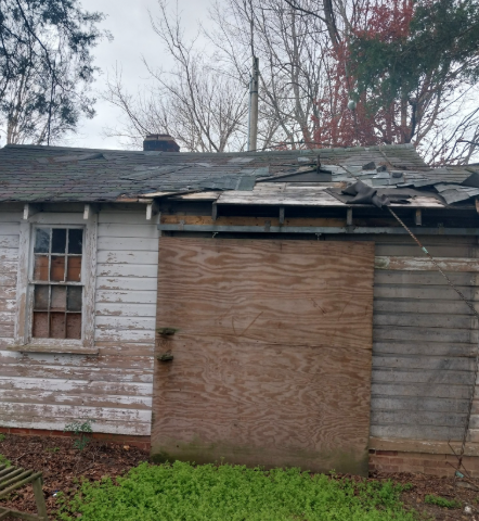 Dilapidated white house with boarded-up door, broken roof, and overgrown yard