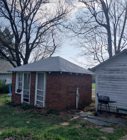 Small brick shed beside a gray garage in a yard with bare trees and patchy grass.