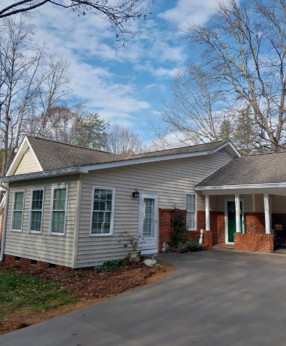 Single-story beige house with white trim, brick porch, and a driveway under a partly cloudy sky.