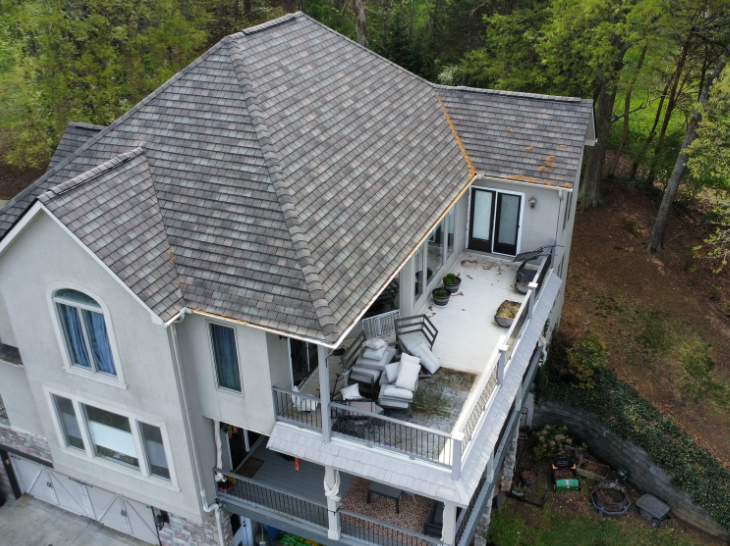 Aerial view of a gray house with a large roof and second-floor balcony surrounded by trees.