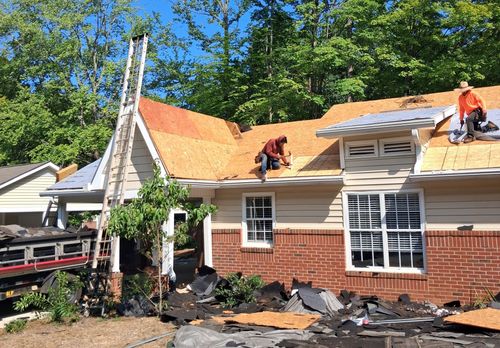 Roofers working on a house roof with orange shingles and brick siding, surrounded by trees.