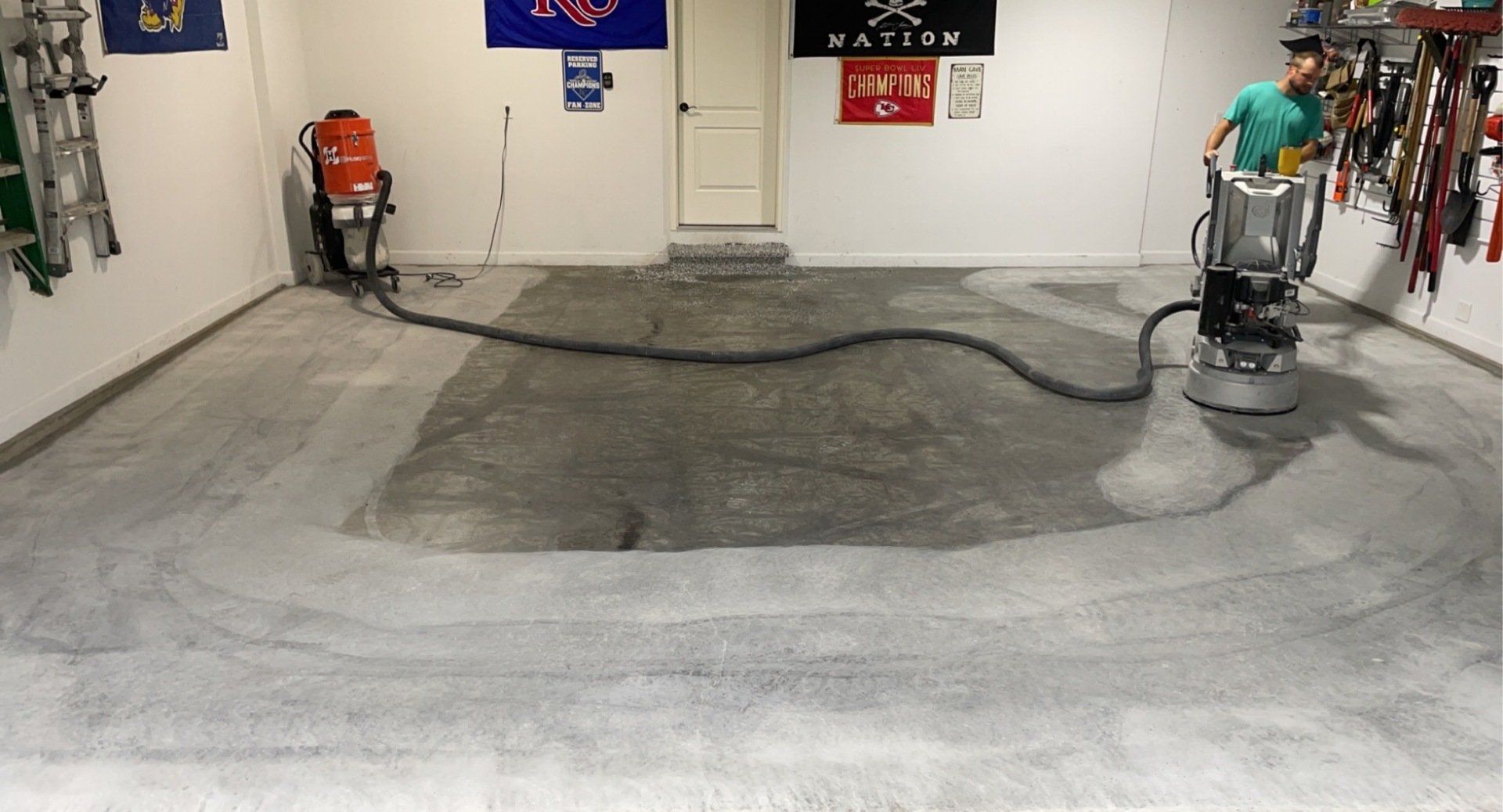 Person operating a concrete floor grinder in a garage. Grey concrete floor, black hose, white walls, tools.