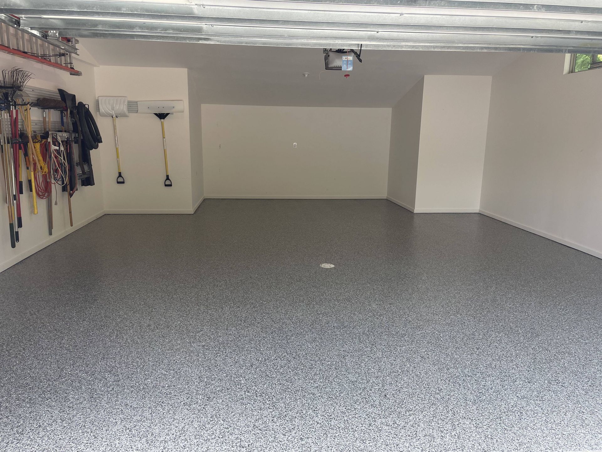 Empty garage with gray speckled floor, white walls, and tools hanging on the left wall.