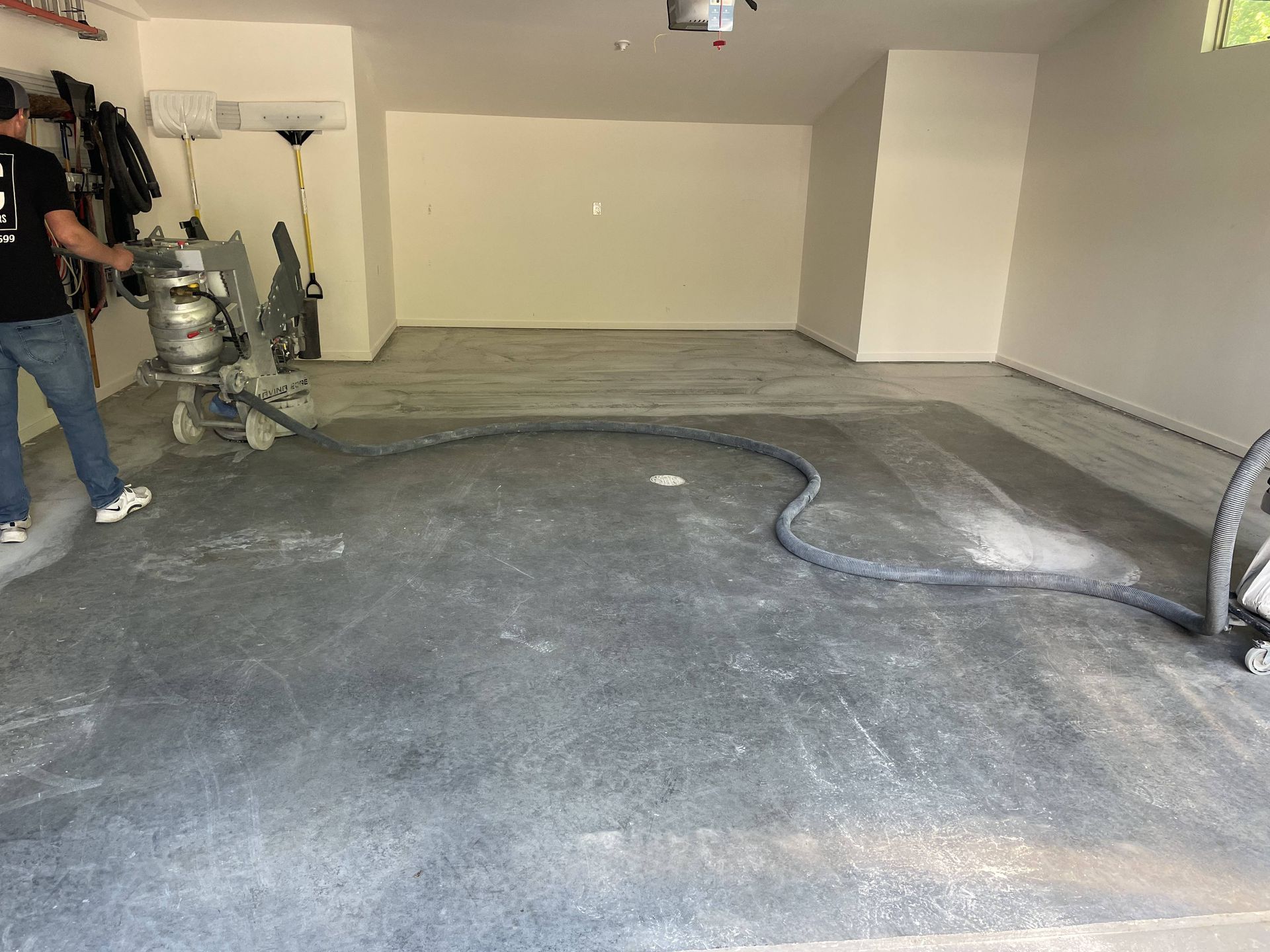 Person grinding a garage floor with a machine. Dust hose snakes across the gray concrete.