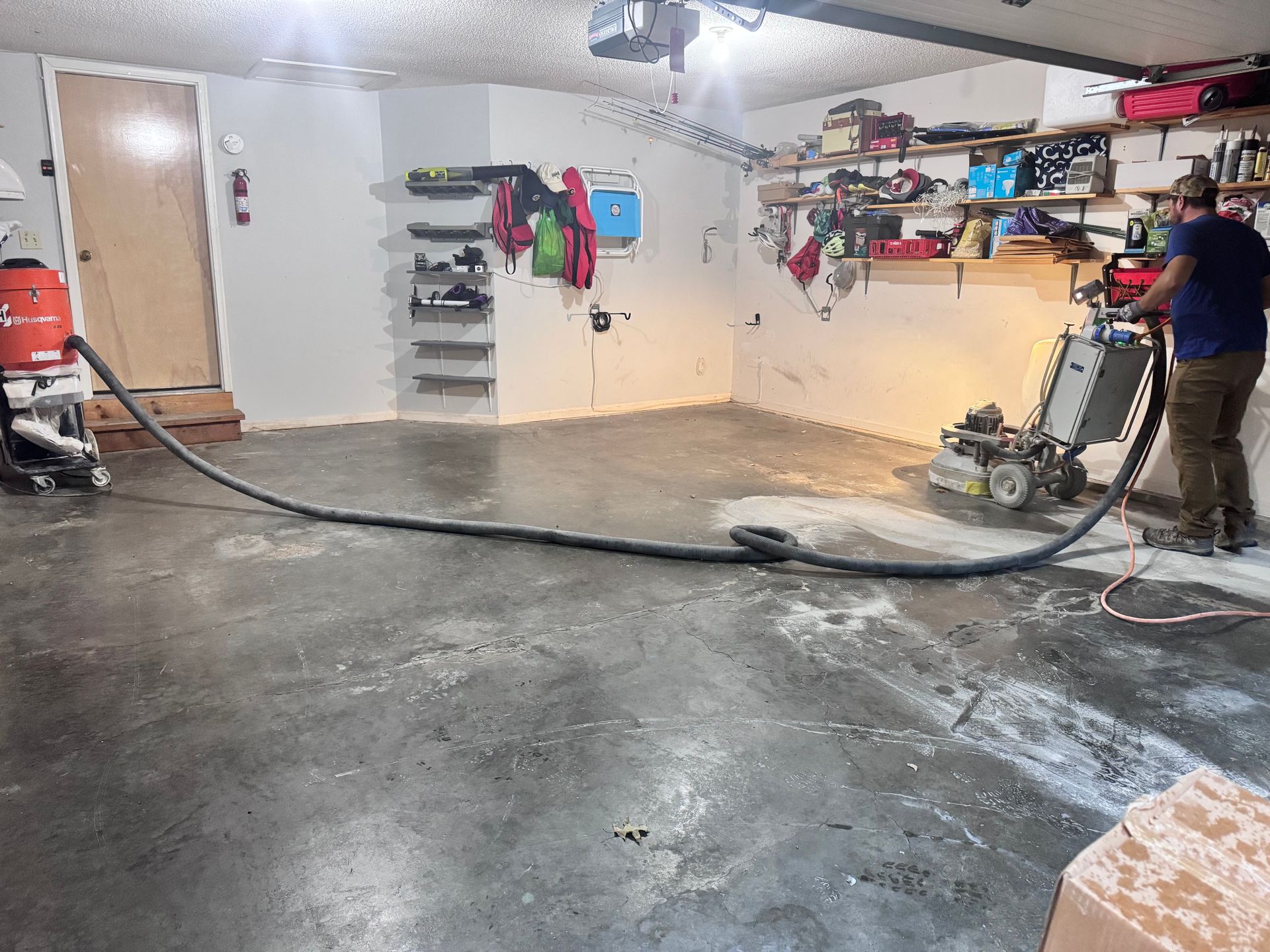 Man using a floor grinder in a garage; concrete floor, dust collection hose.
