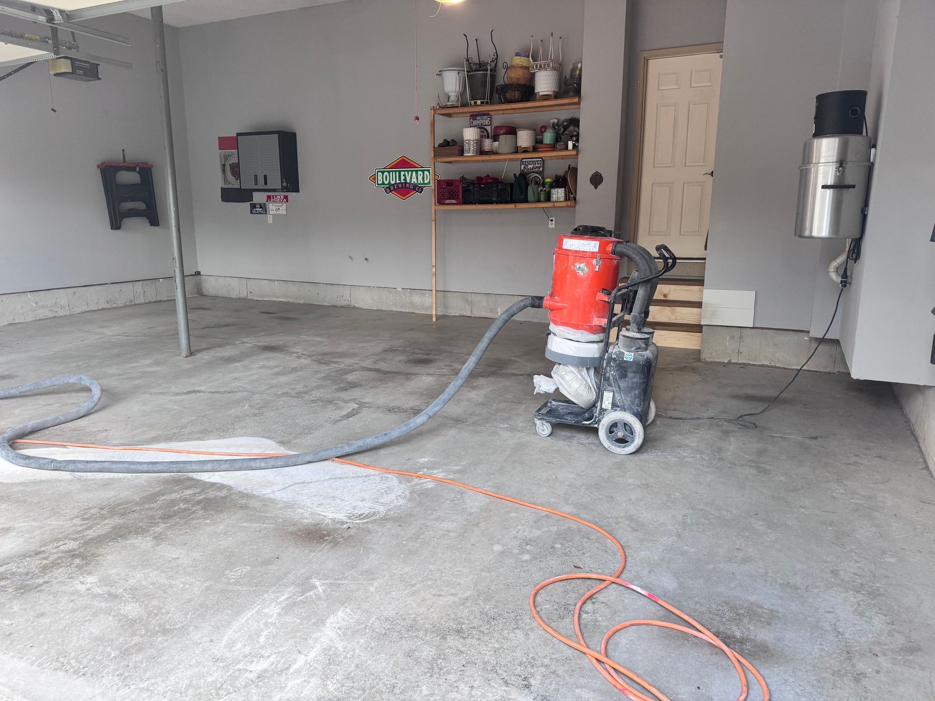A concrete grinder in a garage; gray floor and walls. Orange extension cord and hose are present.