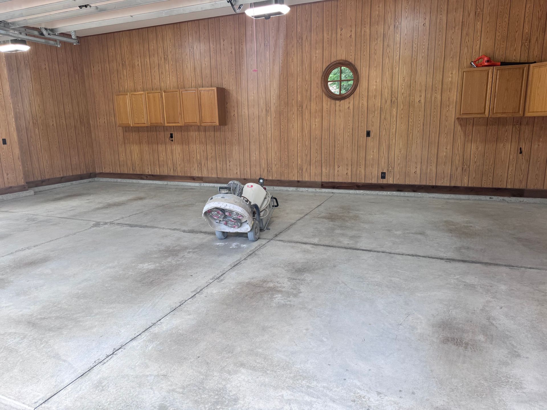 A concrete floor being ground by a machine in a garage with wood paneling and cabinets.