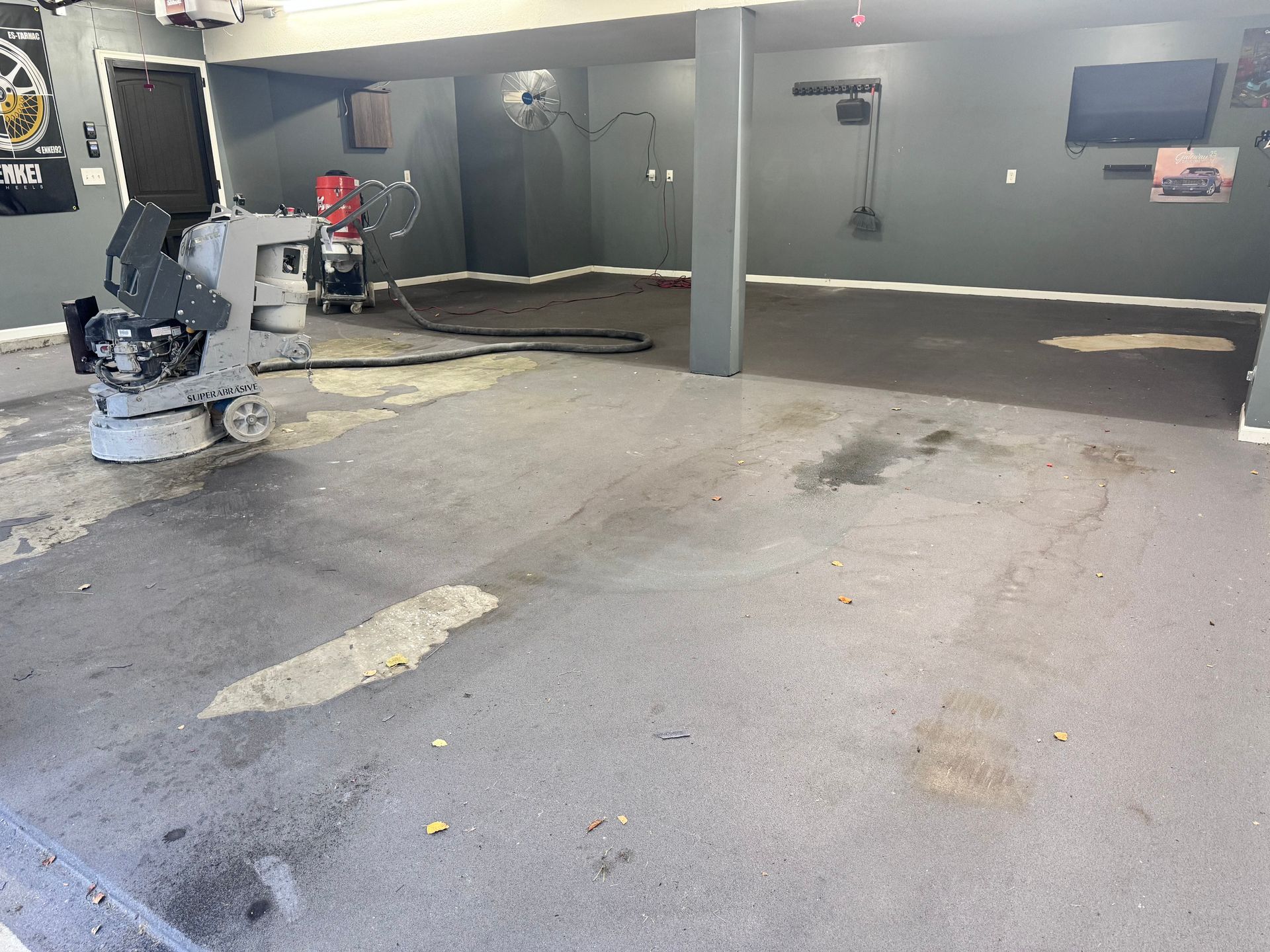 A concrete floor being ground with a machine in a garage; gray walls, equipment, and stains visible.