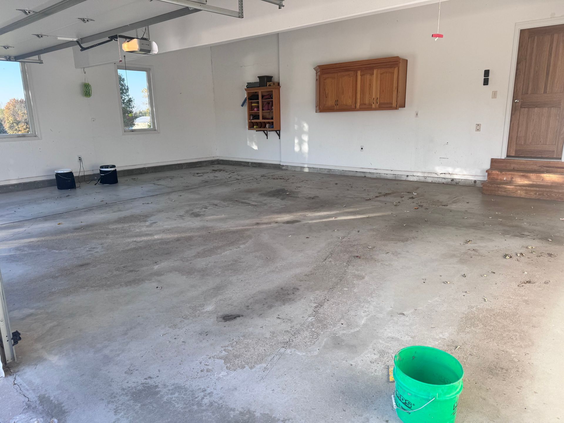 Empty garage with gray concrete floor, white walls, and wooden cabinets. A green bucket sits in the foreground.