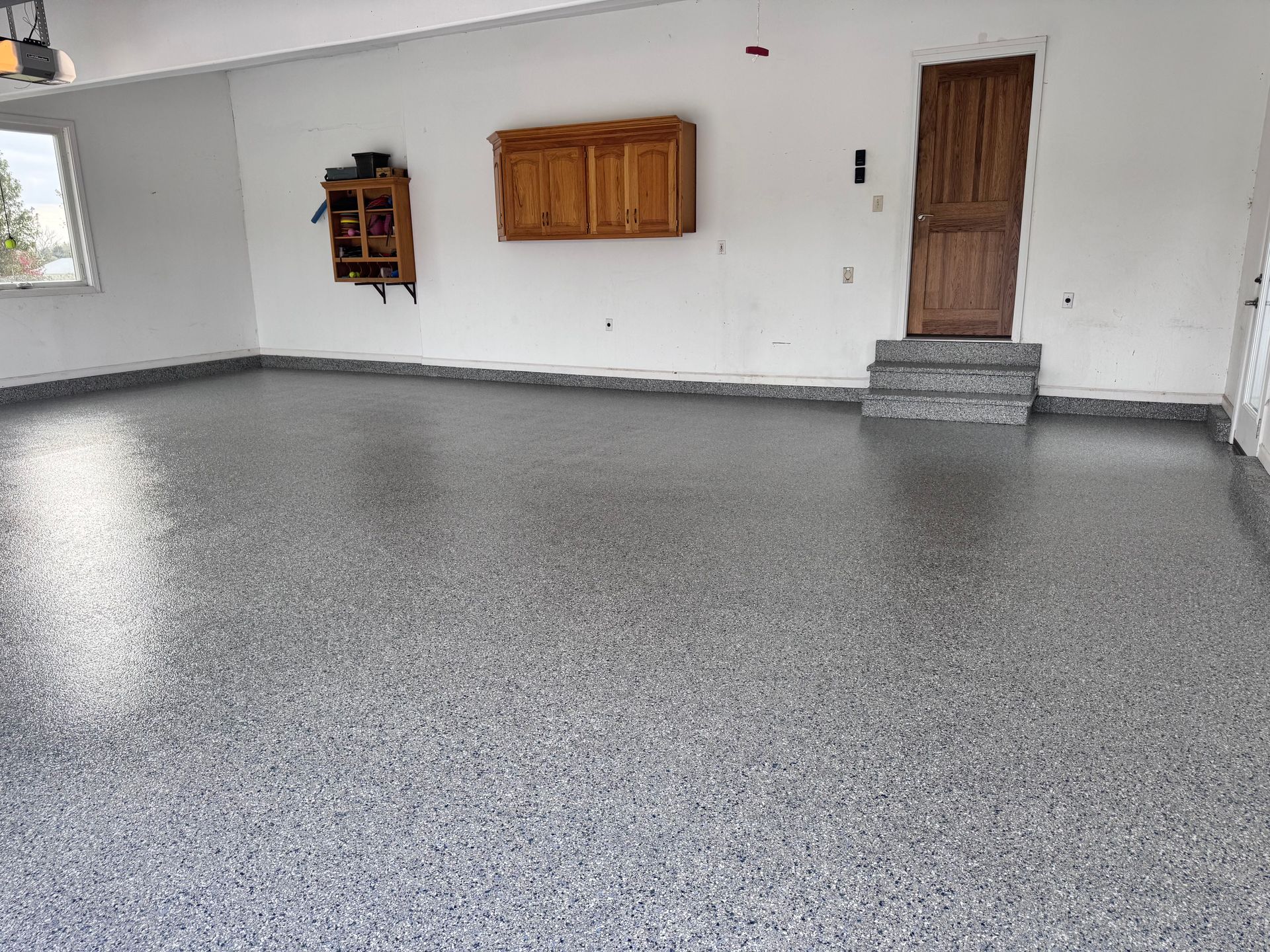 Garage with a gray speckled epoxy floor, cabinets, door, and stairs.