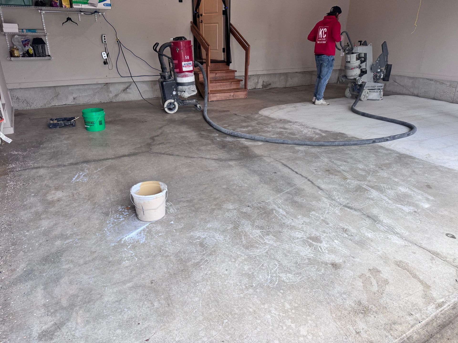Person using machinery to grind concrete floor inside a garage. White dust and equipment visible.