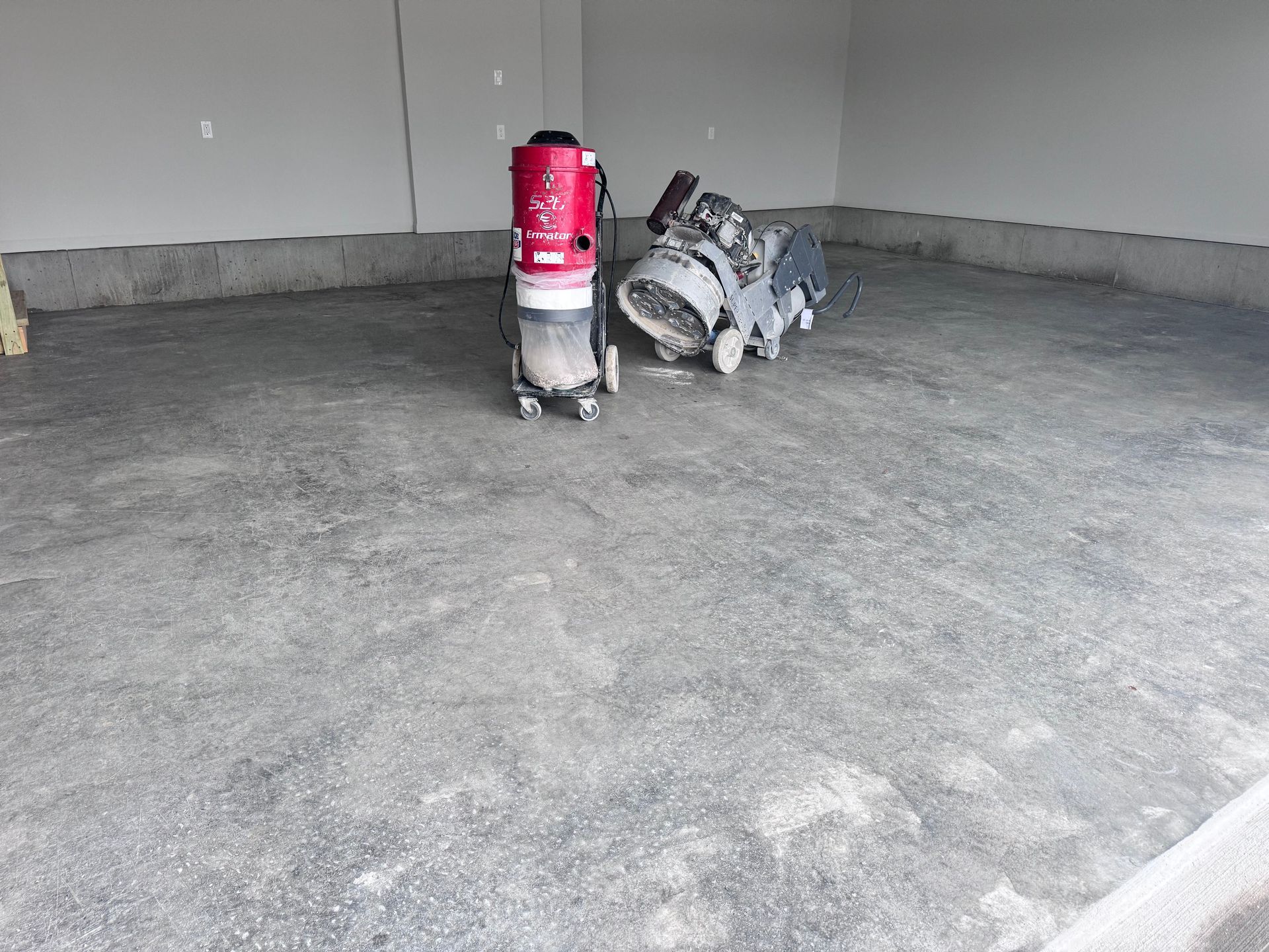 Grinding concrete floor with industrial machinery, red vacuum cleaner, gray walls.