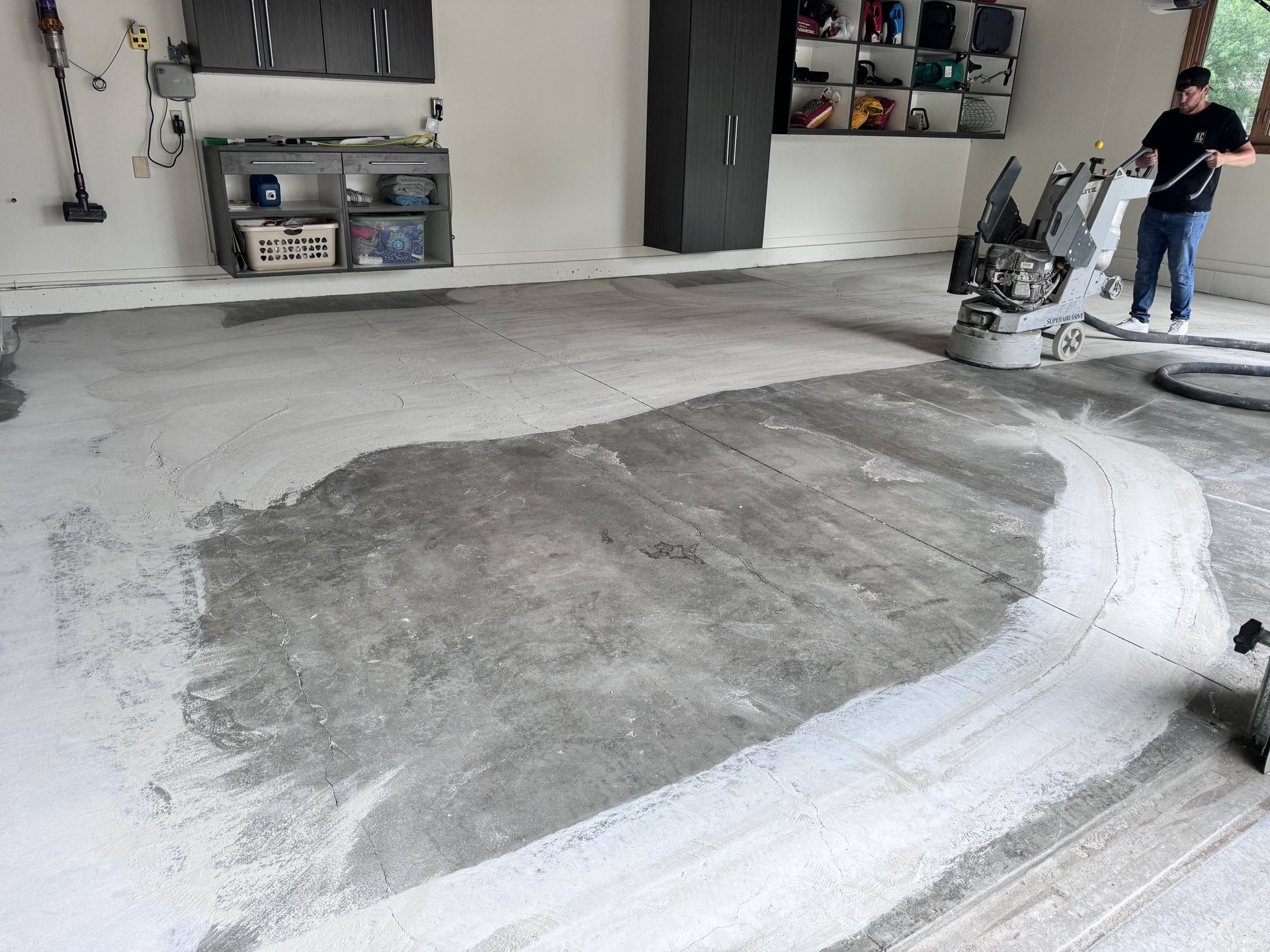 Person operating a floor grinder in a garage; concrete floor partially ground; white dust present.