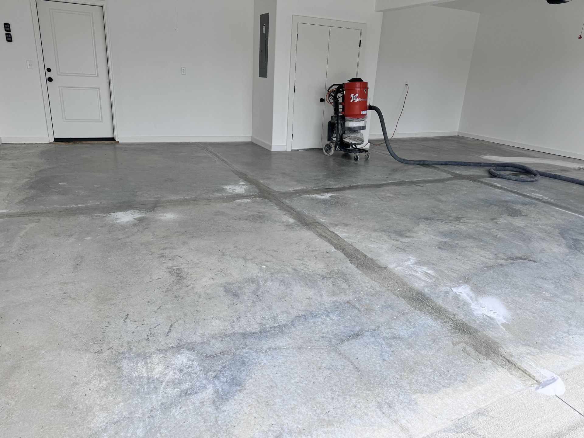 Garage floor being ground by a machine, dust collection hose attached. White walls and doors.