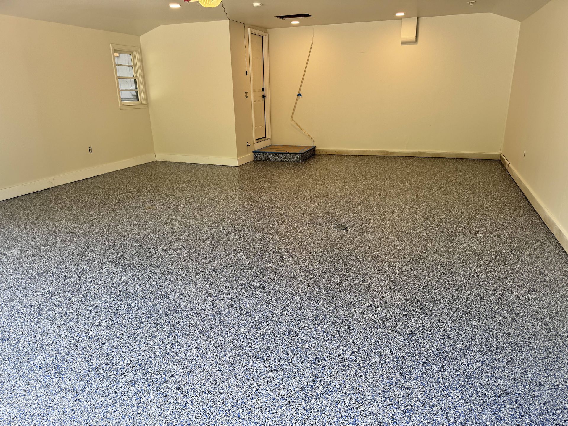 Garage with speckled epoxy floor, beige walls, and a small window. Door in the back leads to a lower level.
