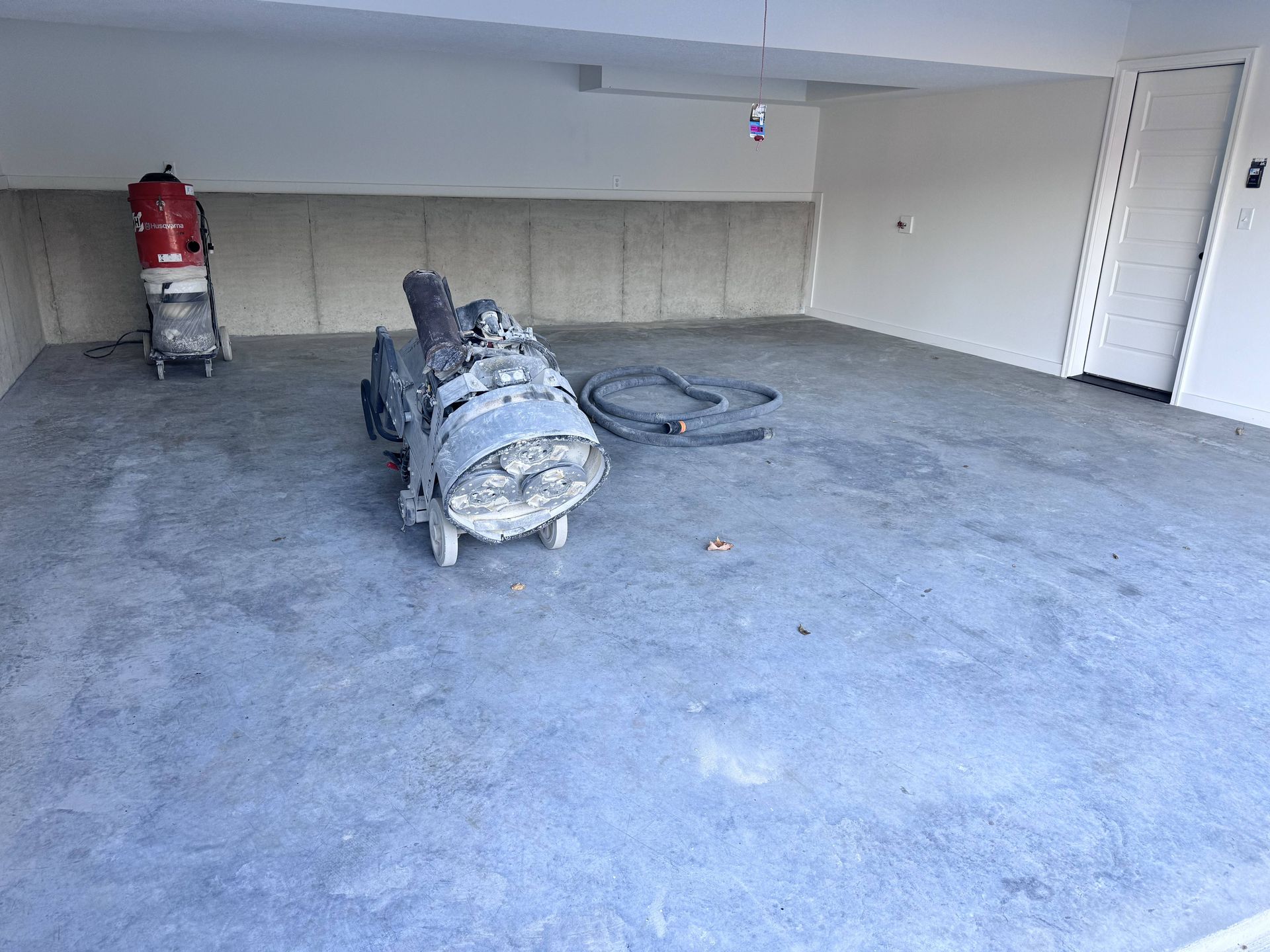 Grinding machine on a gray concrete floor inside a garage. A red vacuum cleaner is in the background.