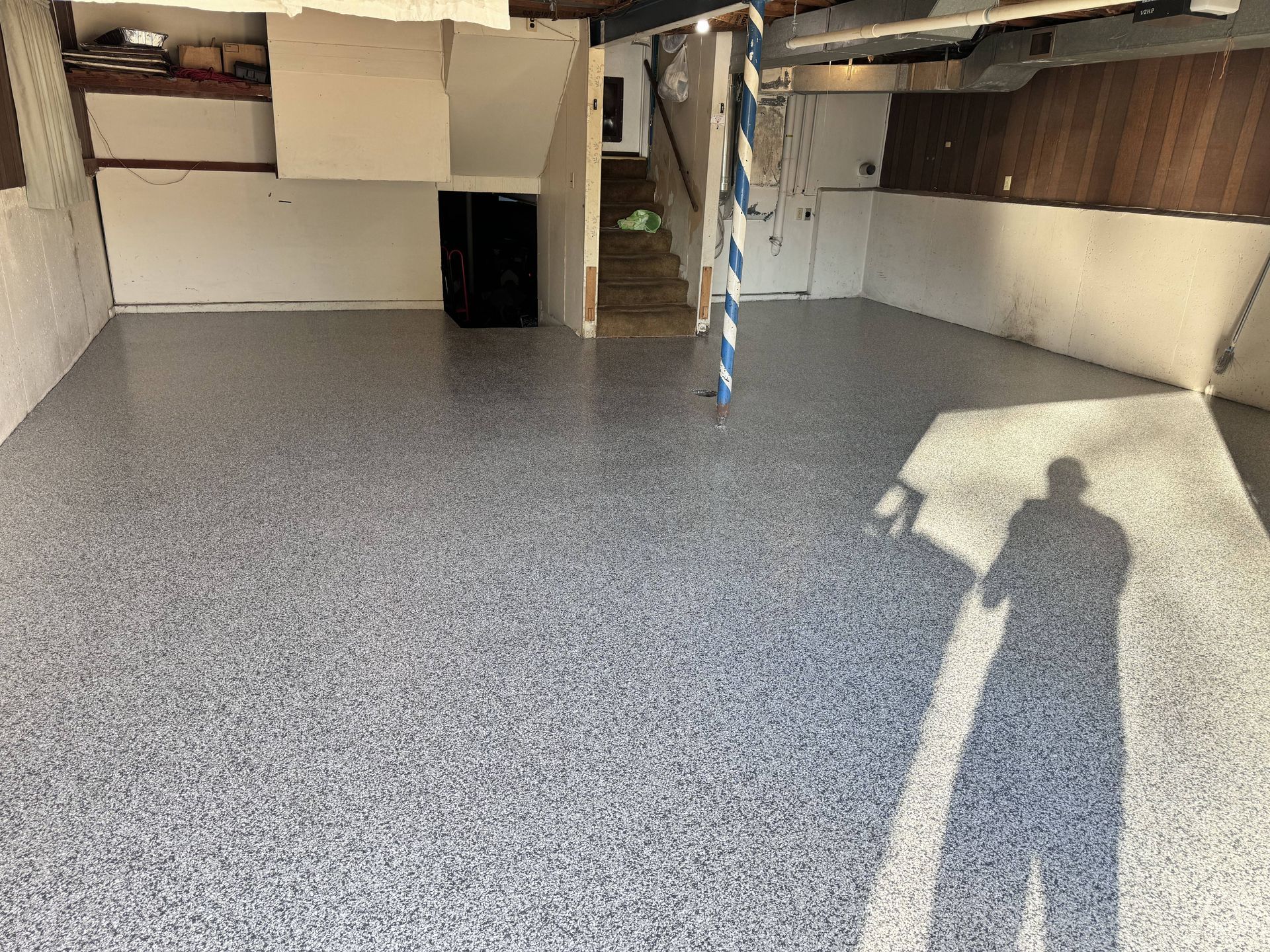 Spacious room with speckled gray floor. White walls and wood paneling. Staircase and shadow of person.
