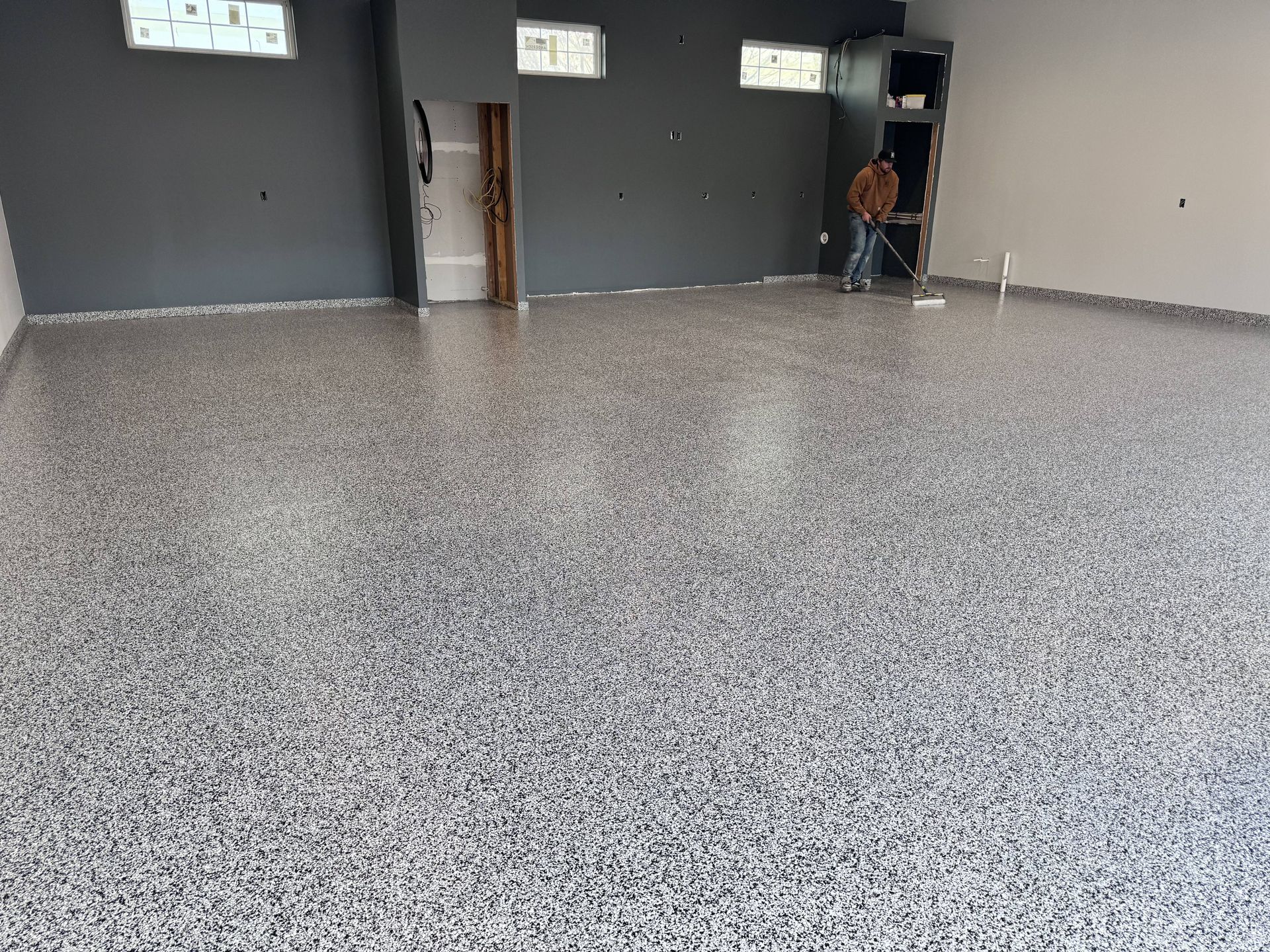 A room with a gray speckled epoxy floor, gray walls, and a person working in the background.