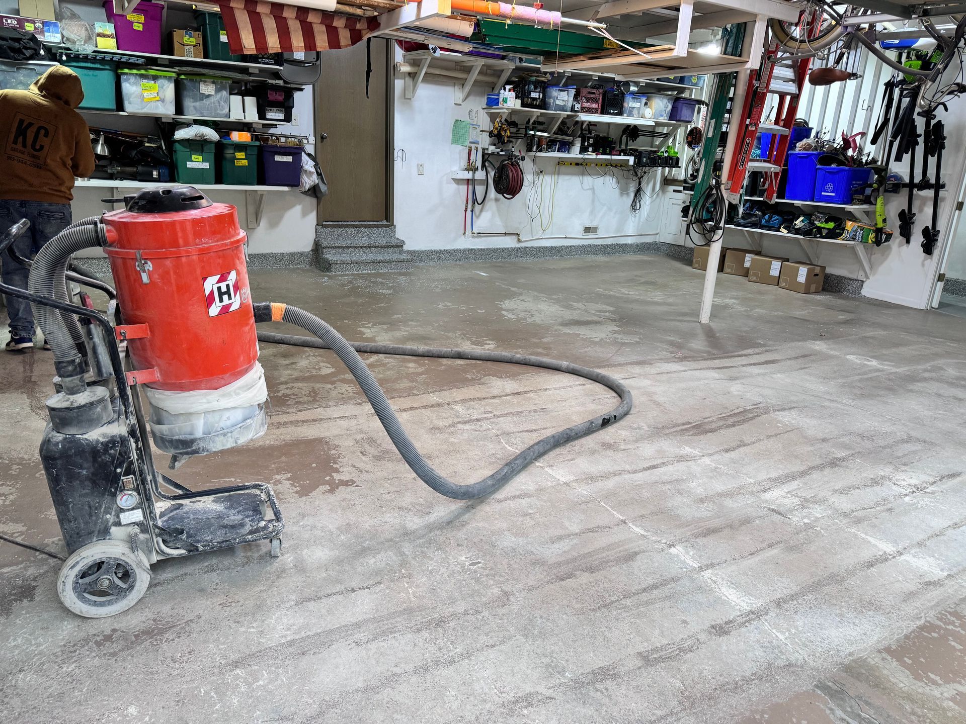 Floor grinder, red and black, in use on a concrete floor inside a garage, next to a person.