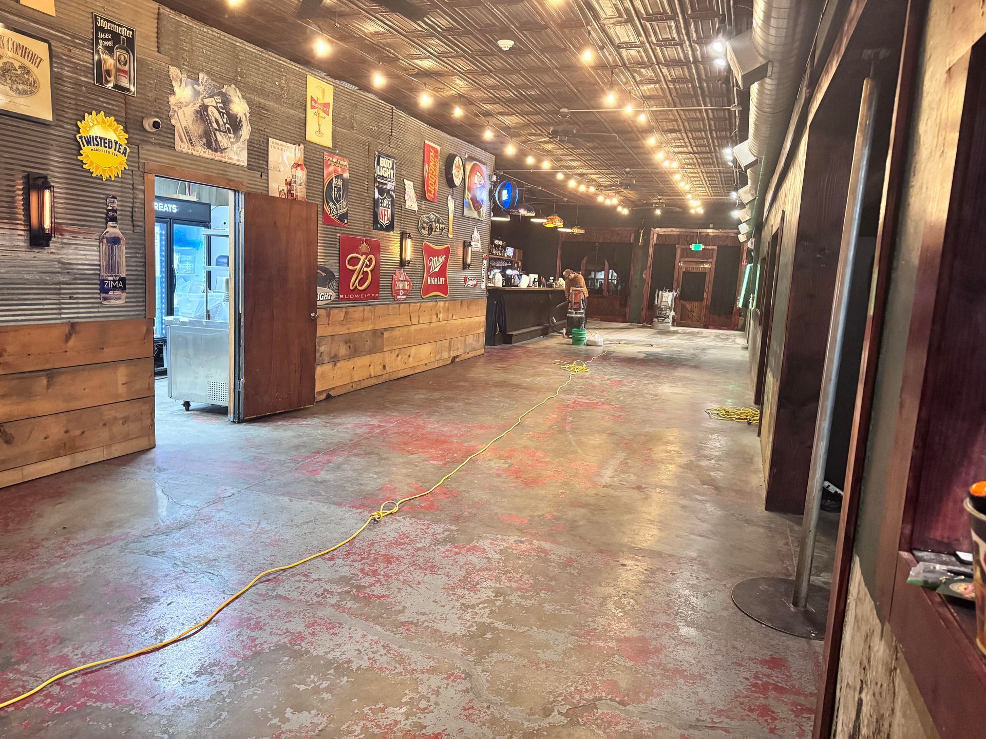 A dimly lit bar interior with a long counter and posters on the wall; string lights overhead.