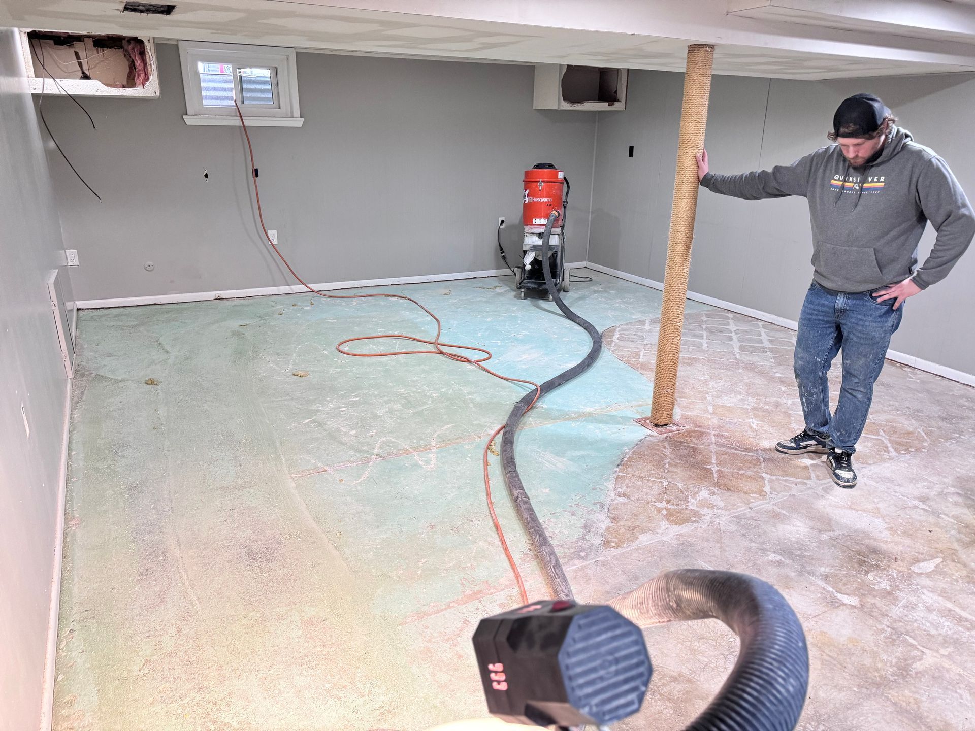 Man standing in unfinished basement, vacuuming insulation; beige walls, exposed beam.