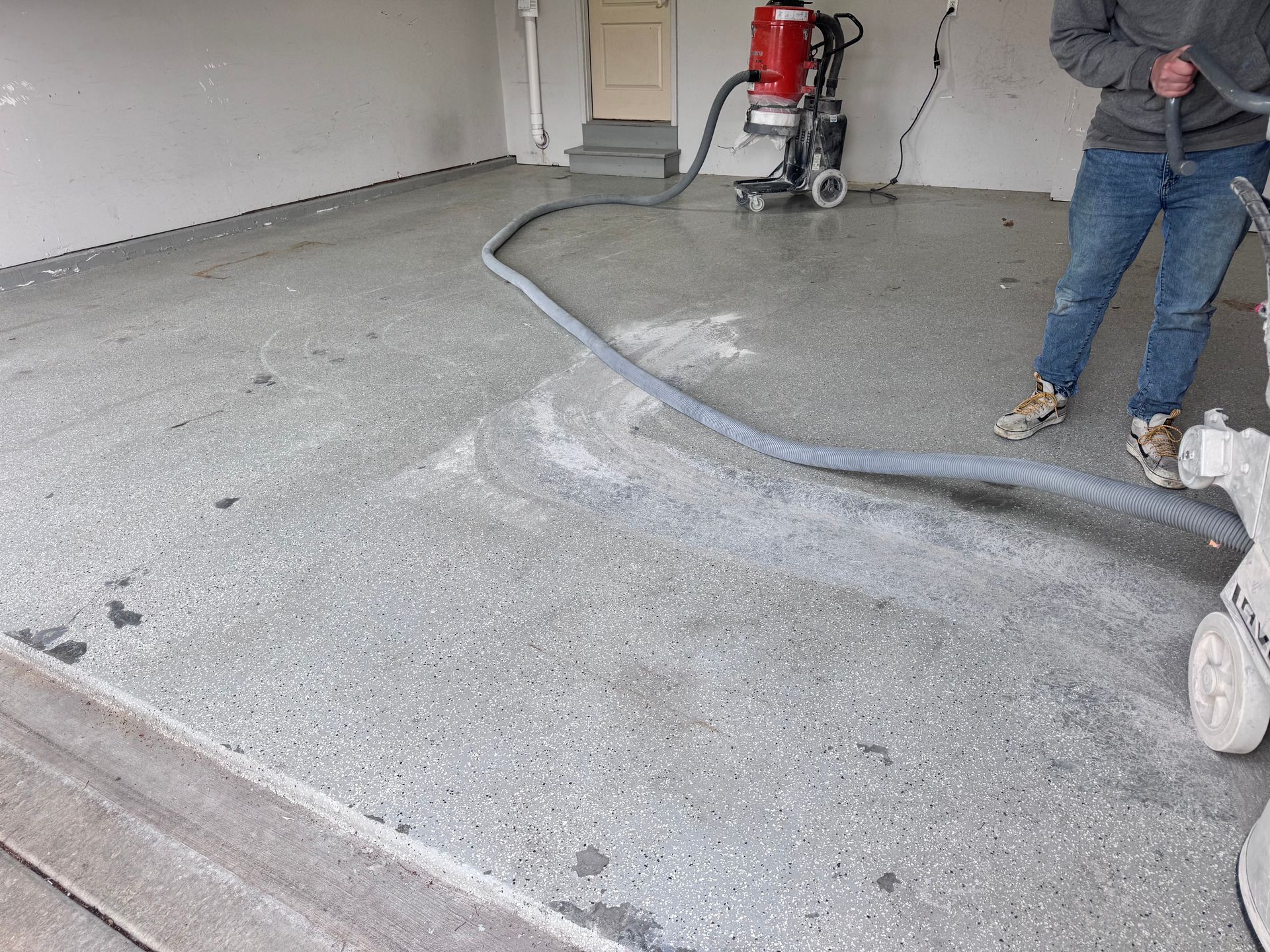 Person using floor grinder in a garage; gray concrete floor being prepared, red machine.