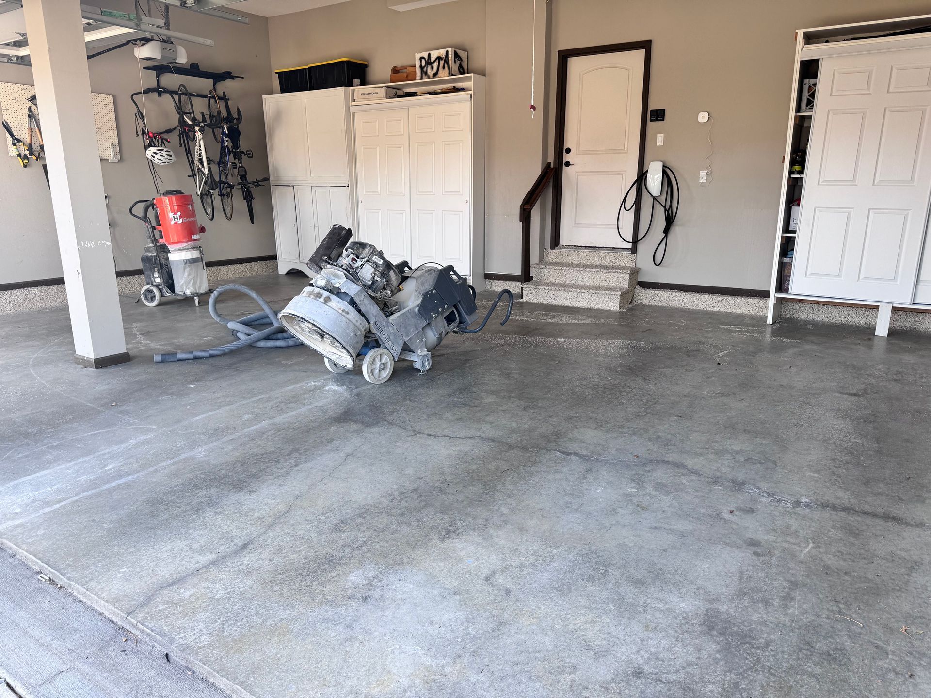 Concrete floor grinding with machinery in a garage. Grey concrete, white cabinets, and a white door are visible.